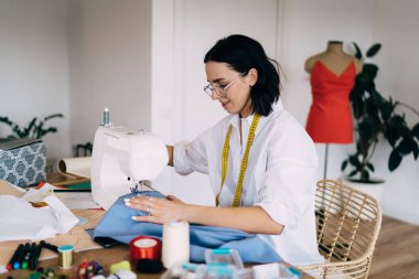 Günlük giysiler ve gözlüklerle atölyedeki masada otururken dikiş makinesinde çalışan etnik zanaatkar kadının yan görünüşü
