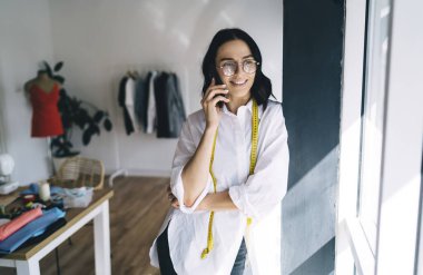 Beyaz bluzlu neşeli genç bayan terzi omuzlarında santimetre ile duvara yaslanıyor ve atölyedeki pencereye bakarken akıllı telefondan konuşuyor.