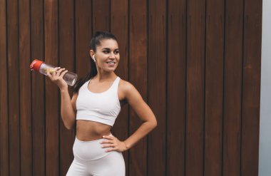 Beyaz atkuyruklu, uzun atkuyruklu, taytlı, TWS kulaklıklı, sırıtan, ahşap duvarda elinde bir şişe suyla dikilmiş duran genç İspanyol sporcu.