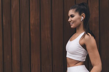 Reklam metni için arka planda duran kablosuz kulaklıklı sağlıklı genç bir kadın. İnce, güzel vücutlu çekici esmer kadın kulaklıkla müzik dinliyor, müzik listesinden zevk alıyor.