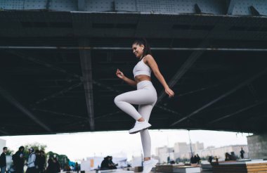 Uzun atkuyruklu sporcu kılıklı pozitif İspanyol bayan sporcunun şehir sokaklarındaki antrenman sırasında köprü altında adım atarken gülümsemesi.