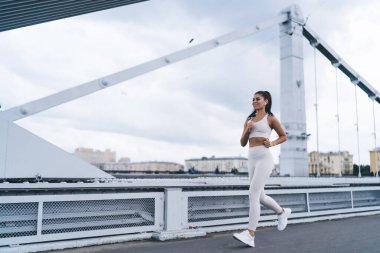 Aktif giyimli, uzun siyah saçlı, kablosuz kulaklıklı ve köprüde koşarken gülümseyen ve müzik dinleyen kararlı genç bayan.