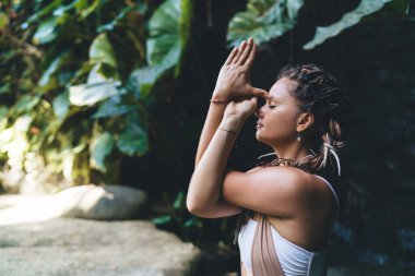 Yeşil tropikal parktaki yoga eğitimi sırasında açık renk kıyafetler giyen sakin kadın Gomukhasana egzersizi yapıyor.