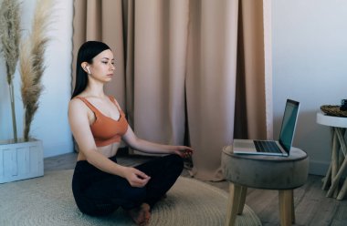 Spor giyimli, TWS kulaklıklı, Lotus 'ta dizüstü bilgisayarın önünde oturmuş, yoga sırasında evde rahat bir odada meditasyon yapan genç bir kadının yan görüntüsü.