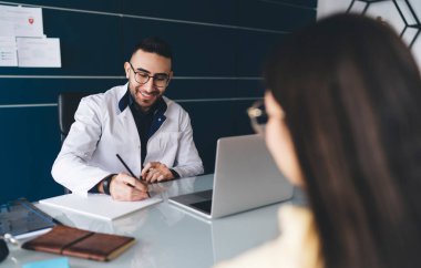 Gözlüklü genç bir erkek doktor, isimsiz mahsul kadın hastasıyla randevusu sırasında reçeteli ilaç alıyor.