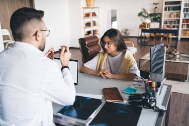 Genç erkek osteopat doktor klinikte oturmuş genç bayan hastayı anlatıyor ve birbirlerine bakarken gerekli omurga ve tedavi modelini gösteriyor.