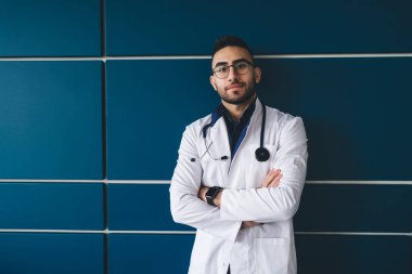 Beyaz cüppeli, omuzlarında steteskop bulunan gözlüklü, kolları katlanmış ve kameraya bakan güvenilir bir etnik erkek pratisyen.
