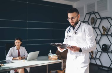 Gözlüklü erkek doktor panoda gazete okurken, kadın meslektaşım da güncel ofiste dizüstü bilgisayarla çalışıyor.