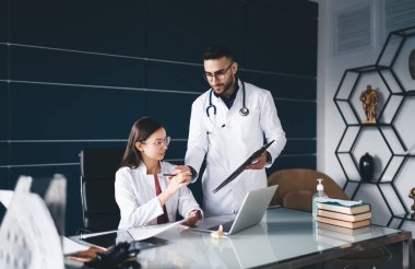 Genç erkek pratisyen hekimin kadın iş arkadaşına kâğıtlı kalem vermesi çağdaş hastane ofisinde çalışma sorunlarını tartışırken.