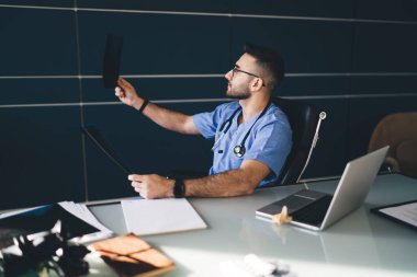 Çağdaş iş yerindeki mavi üniformalı doktorun yan görünüşü ve klinikte sıkı çalışırken röntgen taramasına bakmak