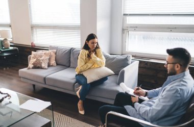 Koltukta oturan yüksek açılı kadın hasta erkek terapiste koltukta oturup notlar yazma sorununu anlatıyor.