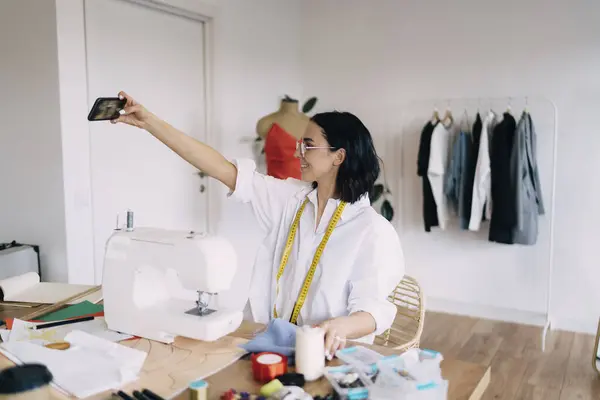 stock image Satisfied ethnic female dressmaker sitting at table with sewing machine and taking self portrait on smartphone while working in studio