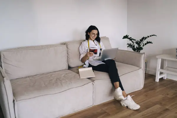 stock image Full body of positive female designer in smart casual outfit sitting on comfortable couch with sketches and working on laptop during workday