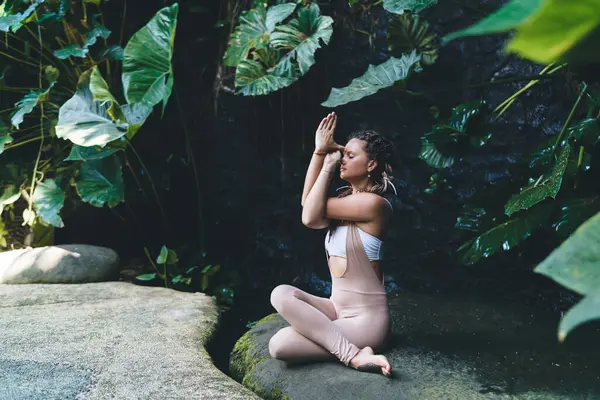 stock image Side view of focused slim female in stylish sportswear performing Gomukhasana while sitting on stone in green tropical park during yoga session