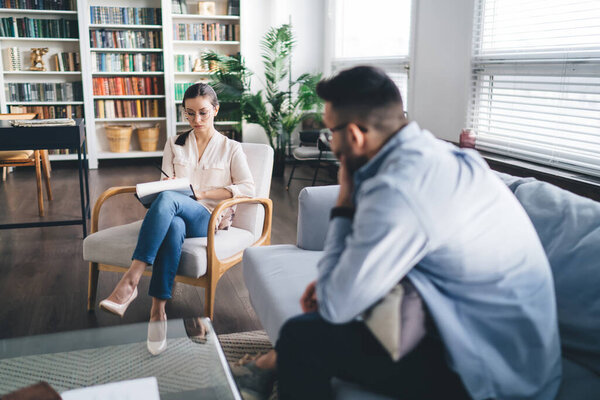 Side view of male patient speaking while psychologist writing down information in notepad and listening to during psychotherapy appointment in modern office