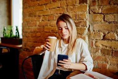 Modern kafeteryada çalışırken cep telefonundan mesaj atan, bir fincan sıcak içecek ile çalışan bir kadın.
