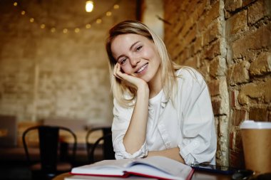 Olumlu genç bayan gülümseyen ve kameraya bakarken masada elinde defter ve kahve bardağıyla otururken başını kaldırıp zaman geçiriyor.