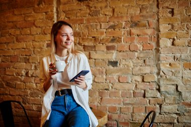 Bir yandan gülümseyen, bir yandan da kafeteryada boş vakitlerin tadını çıkarırken bir yandan da elinde kahve ve cep telefonuyla tuğla duvarın yanındaki masaya bakan genç bir kadın.