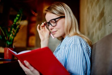 Genç bir kız öğrencinin, elinde gözlüklerle masada oturup bulanık kafede notlar okuyarak gözlüklerini ayarlayışının yan görüntüsü.