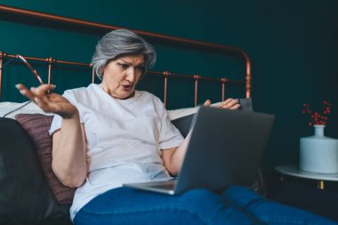 Tatminsiz, gri saçlı, sıradan elbiseli bir kadın yatakta oturup video aracılığıyla konuşuyor ve netbook kullanarak kollarını ayırıyor.