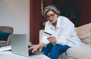 Orta yaşlı kadın şık giyinmiş, gözlüklü, kredi kartı bilgilerini okuyan, dizüstü bilgisayar kullanan ve evdeki oturma odasında internetten sipariş veren.