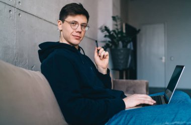 Elinde elektronik sigarayla kanepede oturmuş evdeki oturma odasında dizüstü bilgisayar projesiyle kameraya bakan genç bir adamın yan görüntüsü.