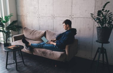 İnternette gezinen, rahat bir kanepede oturup uzaktan proje yapan konsantre genç bir adam.