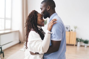 Afro-Amerikalı bir adamın aydınlık odada dikilirken kız arkadaşını kucaklaması ve öpmesi.