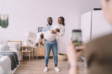 Afro-Amerikalı erkek, evde rahat bir yatak odasında dikilirken ve akıllı telefonlu bulanık bir adam fotoğraf çekerken gülümseyen ve birlikte kameraya bakan kadınları kaldırıyor.