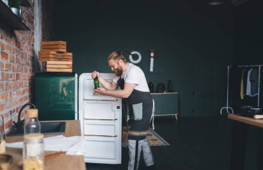 Beyaz tişörtlü ve önlüklü gülümseyen erkek aşçının, evdeki boş buzdolabının önünde dikilirken yeşil cam şişe sergilediği yan manzara.