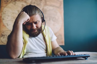 Günlük kıyafetleri ve kulaklıkları olan endişeli sakallı bir adam bilgisayarda, duvarda eski bir haritayla oyun oynarken masada klavyeyle oturuyor.