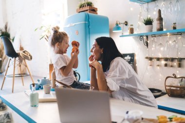 Beyaz tişörtlü mutlu genç annenin modern mutfakta elinde mandalinalarla oturmuş birbirine bakan kızına gülümsemesi.
