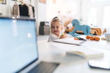 Masada keçeli kalemler ve pastel kalemlerle gülümseyen sevimli kız. Bu arada serbest çalışan anne evde çalışıyor ve ev işi yapıyor.