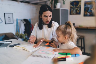Evde çalışan mutsuz, meşgul genç bir kadın, masada oturmuş defterine işaret ederken, çocuk çizim defterinde turuncu kalemle resim çiziyor.