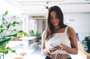 Şık giyinen mutlu kadın ekranlara bakıyor ve cep telefonuyla mesajlaşıyor modern kafeteryada dinlenirken bulanık arka planda.