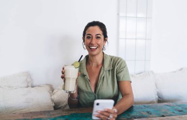 Modern restoranda masada oturmuş margarita kokteylinin tadını çıkarırken şık kıyafetler giyen, gülümseyen ve kameraya bakan mutlu bir kadın.