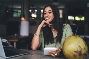 Mutlu genç bir kadın cep telefonu kullanırken ve hafta sonları kafede gülümserken elinde saman ve hindistan cevizi ile masada oturuyor.