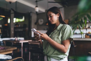 Hafta sonları şehrin modern kafesinde boş vakit geçirirken akıllı telefon kullanan koyu renk saçlı konsantre genç bir kadın.
