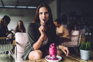 Kafedeki ahşap masada oturmuş chia ve yaban mersinli lezzetli smoothie içerken günlük kıyafetlerini giymiş mutlu kadın kameraya bakıyor.