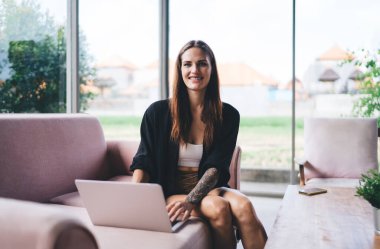 Elinde dövmesi olan, kanepeye oturan ve kameraya bakarken uzaktan bilgisayarla çalışan pozitif genç bir bayan serbest yazar.