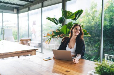 Uzun saçlı ve kolları dövmeli neşeli genç bir kadın çevrimiçi derste tropikal bitkilerin keyfini çıkarırken bilgisayarın ve akıllı telefonun yanındaki ahşap masada oturuyor.