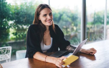 Kolunda dövmesi olan, cep telefonu kullanırken kameraya bakıp gülümseyen ve kafede dizüstü bilgisayarla çalışan genç bir kadın serbest yazar.