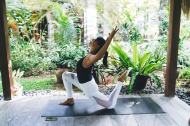 Tahta terasta yoga yapan balerin kızın yan görüntüsü. Uyum ve akıl sağlığı kavramı. Genç esmer atletik kadın spor giysisi giyiyor ve spor minderinde çıplak ayakla dolaşıyor. Bali adası. Güneşli bir gün