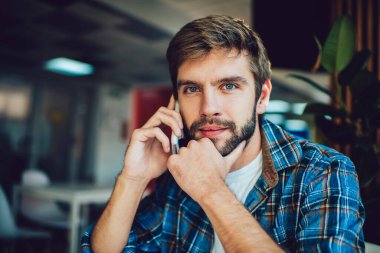 Kareli gömlekli memnun bir erkek kameraya bakarken elleriyle çeneye dokunuyor ve gündüzleri bulanık kafede otururken telefonla konuşuyor.