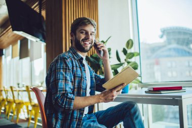 Gülümseyen sakallı erkek tasarımcının elinde taslak varken cep telefonuyla telefon görüşmesi yapması.