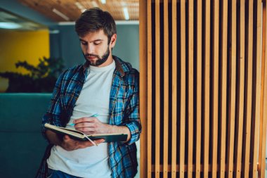 Düşük açılı, günlük giysiler giyen, not defterine notlar yazan erkek çalışanın geniş bir ofiste işe başlaması ve çalışması.