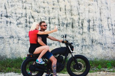 Genç aşıklar özçekim için poz veriyor motosiklet üzerinde oturuyor yaz gezisi yapıyor, çekici sarışın hippi kız sakallı motosikletli sevgilisiyle fotoğraf çekiyor güneş gözlüğü takıyor.