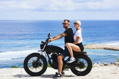 Genç erkek ve dişi bisikletçiler, okyanus kıyısında motosiklet sürerken birlikte seyahat ediyorlar. Yaz tatillerinin tadını çıkarıyorlar. Sıcak yaz gününde motosiklet sürerken.