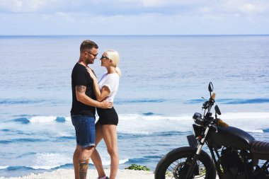 Deniz kıyısında buluşan sevimli çift hafta sonları birlikte olmanın keyfini çıkarıyor, güneş gözlüklü ve modaya uyan aşıklar güzel okyanus sahillerinde motosikletle gezerken sarılıyorlar.