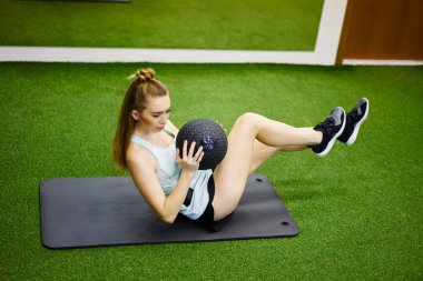 Yukarıdan bakıldığında, spor kıyafetli, mindere uzanmış ve bacaklarını esnetirken karın egzersizi yapan spor salonundaki paspaslı genç bir kadın görülüyor.